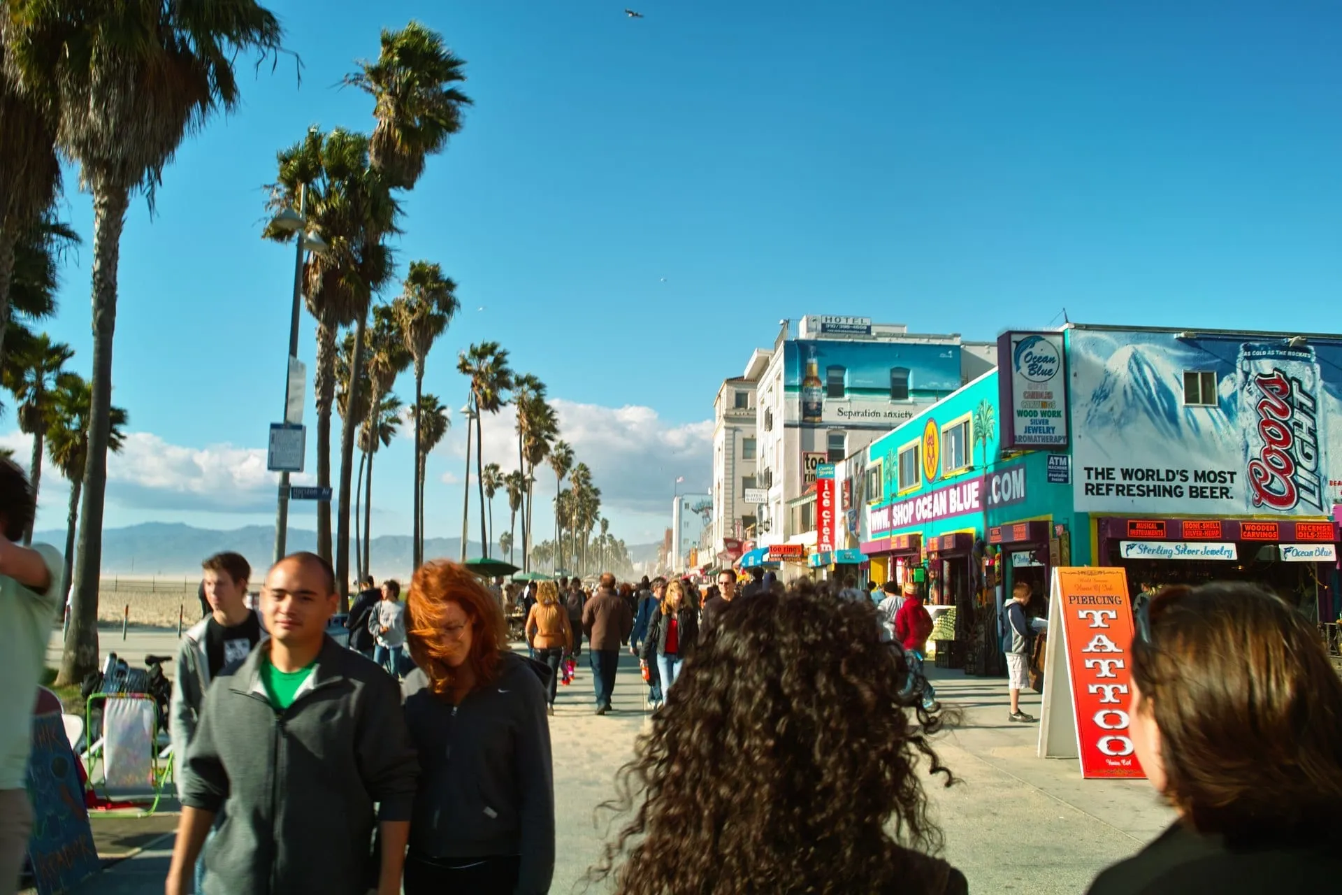 Passanten schlendern über die Promenade von Venice Beach, gesäumt von Palmen und bunten Läden.